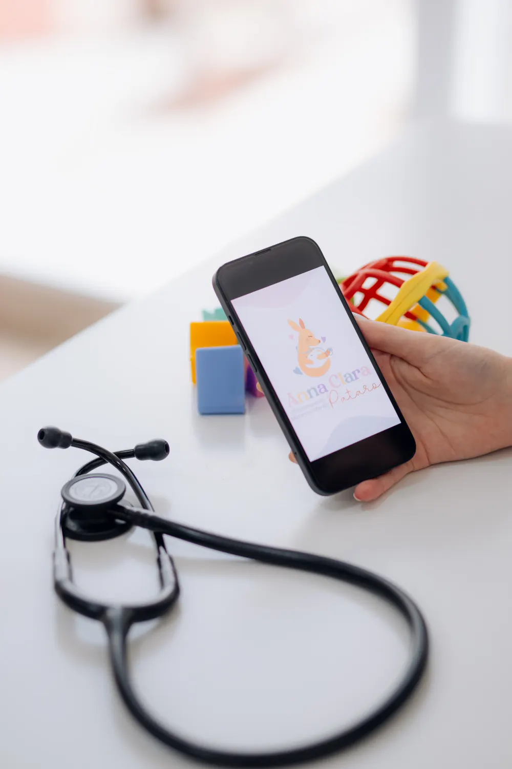 Celular com a marca da Anna Pataro Fisioterapia Infantil, ao lado de brinquedos e estetoscópio
